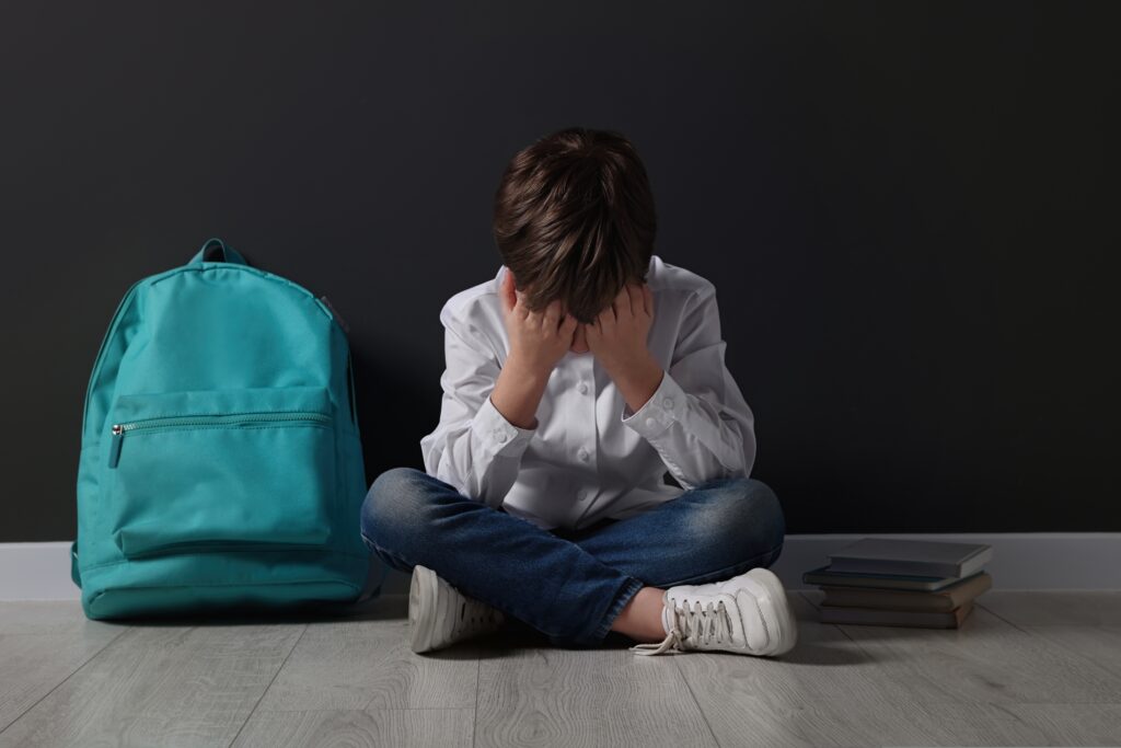 Upset,Boy,With,Backpack,Sitting,On,Floor,Near,Black,Wall.